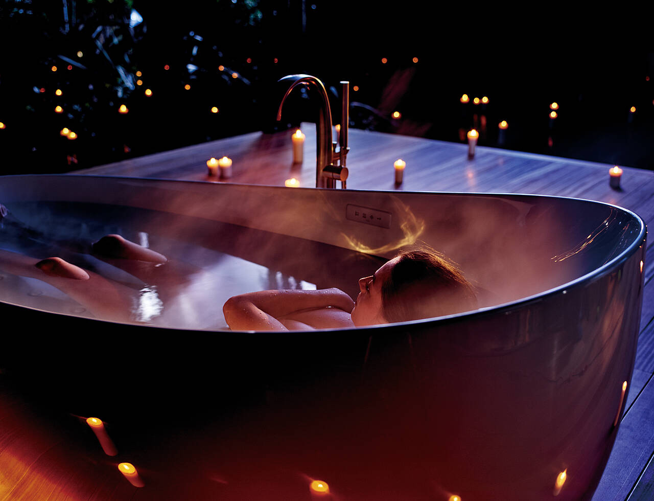 A woman lies in an oval bathtub. Everything except her knees, head and arms crossed over her chest is covered by water. There are candles in the background.