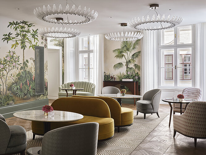 The picture presents a bright and airy lounge interior with large windows, accentuated by elegant furniture in shades of green and gold and two eye-catching chandeliers. The walls are decorated with large murals of tropical plants, which, together with the real plants in pots, give the room a fresh and lively atmosphere.