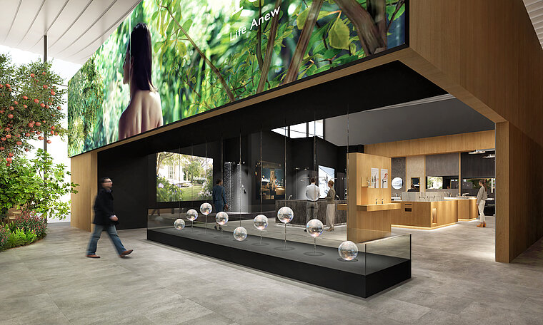 Das Bild zeigt eine moderne Ausstellungshalle mit einem dunkel gefärbten, offenen Stand von TOTO, in dem Dusch- und Armaturenprodukte präsentiert werden, begleitet von klaren Kugeldisplays auf einer langen, schwarzen Theke. Über dem Stand ist ein großes Bildschirmdisplay mit Naturbildern zu sehen, während links im Bild eine grüne Pflanzenwand die natürliche und umweltbewusste Ausrichtung der Marke betont.