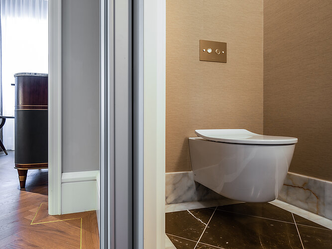 The photo shows a view of a modern, wall-hung toilet in an elegant bathroom with beige-colored walls and a dark patterned marble floor. A luxurious, dark wooden piece of furniture can be partially seen on the left of the picture, and there is a stylish flush control panel on the wall above the toilet.