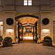 L'image montre l'entrée d'un élégant hôtel illuminée de manière festive, décorée de couronnes et de guirlandes lumineuses. Des lanternes et des bacs à plantes décorés de lumières flanquent le chemin qui mène aux portes vitrées automatiques, conférant à l'entrée une atmosphère accueillante et festive.
