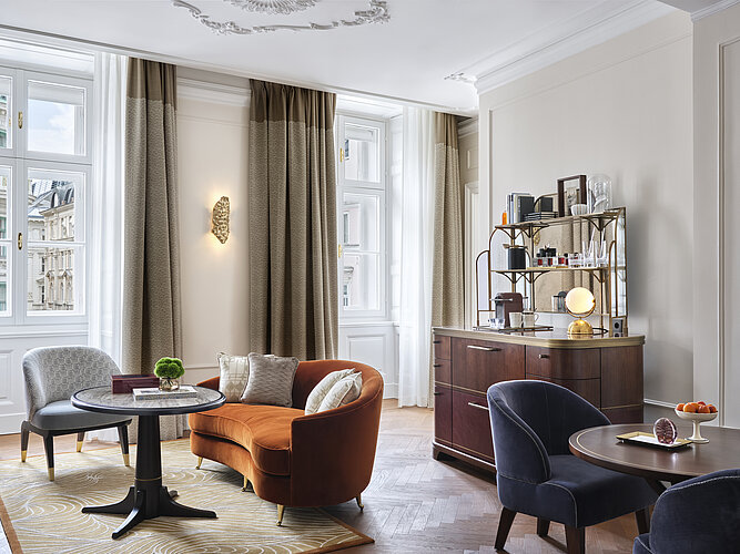 The picture shows a stylishly furnished living room with classic stucco ceilings and large windows that let in natural light. In the room there is an orange armchair, two blue chairs around a round table and an elegant wooden console with a mirror and decorative objects on it.