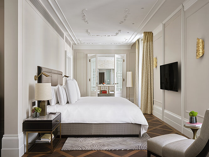 The photo shows a luxurious bedroom with a large double bed in the center, flanked by bedside tables with lamps and opposite a wall-mounted television. The room is characterized by high ceilings with stucco decorations, floor-length curtains and a view through an open door into another room with mirrored walls, which emphasizes the elegance and spaciousness of the room.