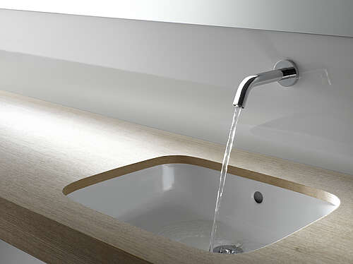 A tap is mounted on a wall above a washbasin. The tap is switched on and a jet of water is coming out of it. 