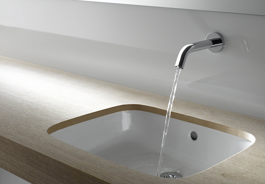 DLE105ANE4_autofaucets Un rubinetto è montato a parete sopra un lavabo. Il rubinetto è aperto e da esso esce un getto d'acqua.